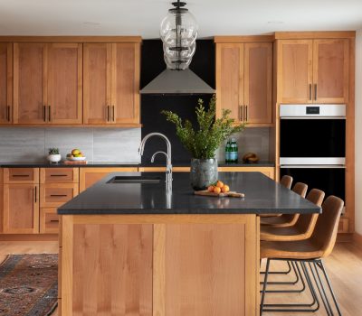 Modern kitchen with wooden cabinetry, black countertop island, and contemporary lighting. Bar stools line the island. Fruit and plant decor add warmth.