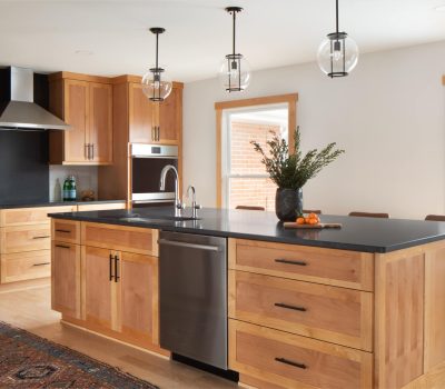 A modern kitchen with wooden cabinets, a black countertop island, pendant lights, and a decorative plant. A colorful rug accents the space.