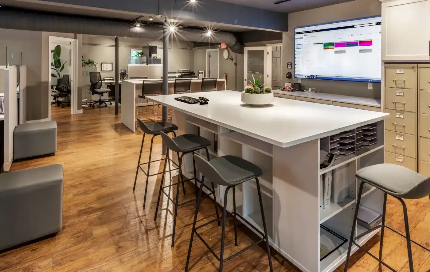Modern office with wooden floors, meeting table, chairs, and computers. Open layout features plants, organized spaces, and a large wall-mounted screen.