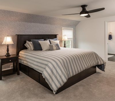 A cozy bedroom features a striped bedding, dark wooden furniture, two lamps, a ceiling fan, and a view of the outside.