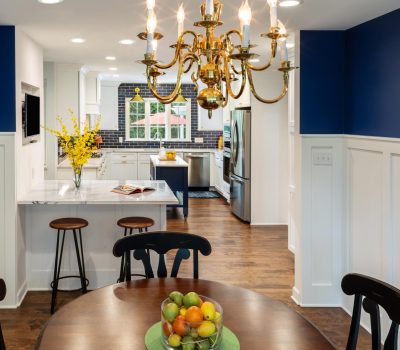 Bright kitchen with wooden floors, blue and white walls, a chandelier, and a dining table. A vase with yellow flowers decorates the counter.