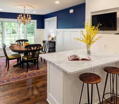Bright dining room features a wooden table, rug, chandelier, and large windows. A kitchen counter with stools and yellow flowers nearby.