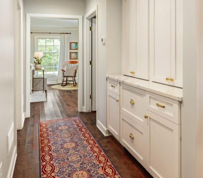 A hallway features a colorful carpet, modern cabinetry, and leads to a bright room with a rocking chair and table with lamp.