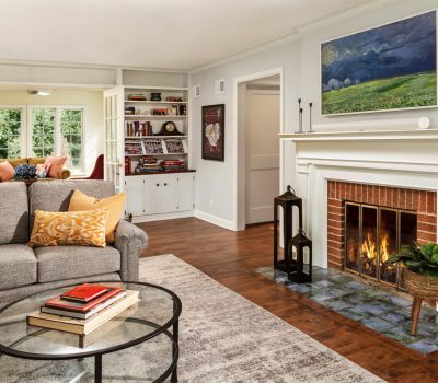 A cozy living room features a fireplace, gray sofa, glass coffee table, art, bookshelves, and natural light from large windows.
