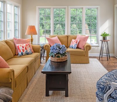 Bright living room with large windows, yellow sofas, colorful pillows, and a stylish coffee table centerpiece. Wood floor and neutral rug complete the decor.