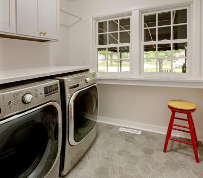 Bright laundry room with front-loading washer, dryer, red stool, modern hexagonal tile floor, and large windows offering a view of a suburban street.