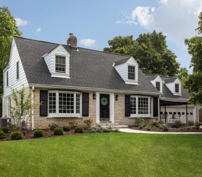 Charming two-story house with dormer windows, stone and siding exterior. Lush green lawn, trees, and decorative wreath on front door. Peaceful suburban setting.