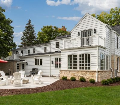 A spacious white house with a stone base, featuring a patio with chairs, fire pit, and red umbrella, surrounded by trees.