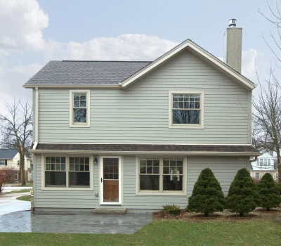 A grey two-story house with a chimney, surrounded by small trees and a concrete driveway, under a cloudy sky.