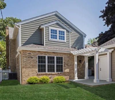 A two-story suburban house with brick and siding exterior, white pergola, and a neatly landscaped yard featuring a white fence and green lawn.