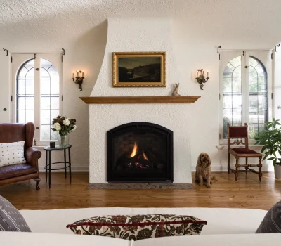Cozy living room features a lit fireplace, leather armchair, arched windows, plant, table with flowers, and a dog sitting on wooden floor.