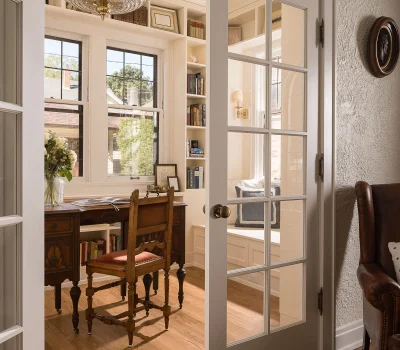 A cozy home office with a wooden desk, bookshelf, chandelier, and French doors, illuminated by natural light from a large window.