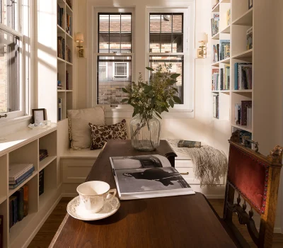 Cozy home library with wooden desk, bookshelves, window seat, and decorative vase on table. Cup and book add to the inviting atmosphere.