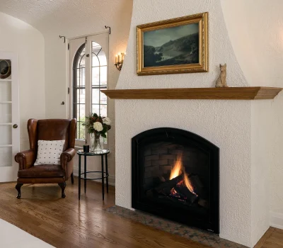 Cozy room with a lit fireplace, brown leather chair, window, framed painting, and a small table with flowers. Warm, inviting atmosphere.