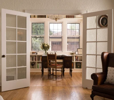 Cozy study room with elegant vintage desk, chair, bookshelf, and French doors, featuring a soft armchair and chandelier on wooden flooring.