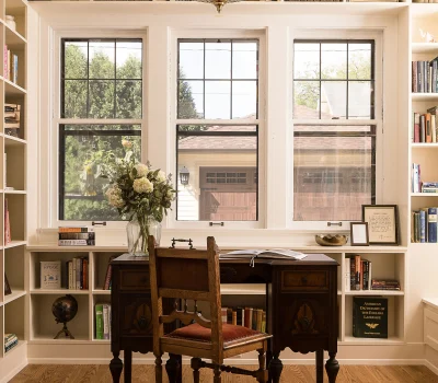 A cozy home office with a vintage desk, wooden chair, chandelier, and bookshelves. Large windows illuminate flowers and a globe.