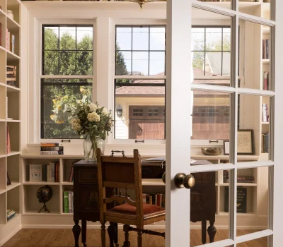 Cozy home office with wooden desk, chair, bookshelves, and a globe. Large windows, chandelier, and bouquet create a warm, inviting atmosphere.
