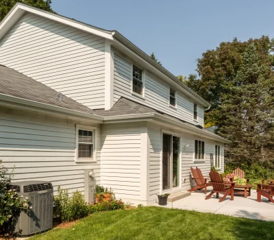 A suburban white-sided house with a spacious backyard, patio, and wooden chairs surrounded by lush greenery under a clear blue sky.