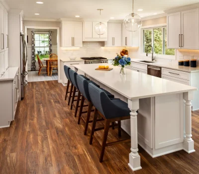 Spacious modern kitchen with white cabinets, wooden floors, island with blue chairs, pendant lights, and a dining area in the background.