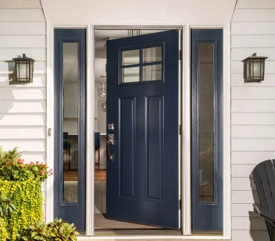 A blue front door partially opens, framed by two glass panels. Flanking plants add greenery. Exterior lights are mounted on white siding.
