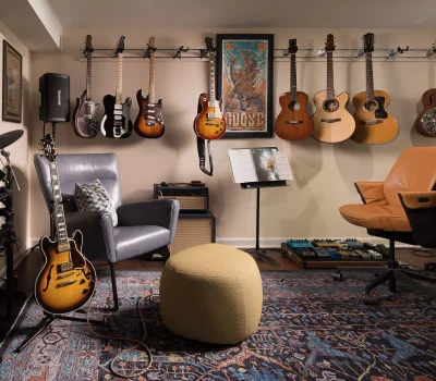Music studio with various guitars on the wall, electronic drum set, amplifiers, sheet music stand, gray armchair, and textured ottoman.