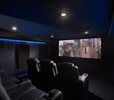 A home theater features plush recliners and muted lighting. The screen displays a classic film scene with two elegantly dressed people conversing outdoors.