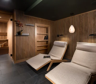 A cozy, modern relaxation room with wooden lounge chairs, wall paneling, a pendant light, and an adjoining sauna featuring a wooden bucket.