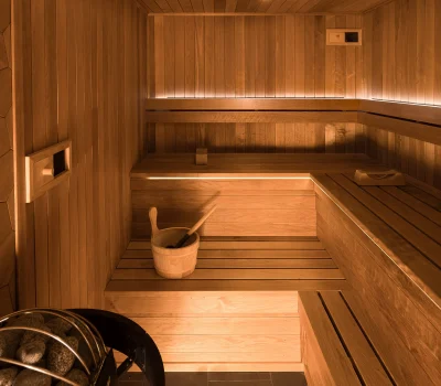 A modern wooden sauna interior features warm lighting, benches, and a stone heater with a bucket and ladle for water. Calm and inviting.