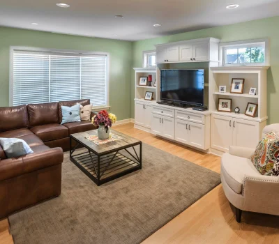 Cozy living room with a brown sectional sofa, armchair, TV on white cabinets, and decorative shelves, highlighted by soft green walls.