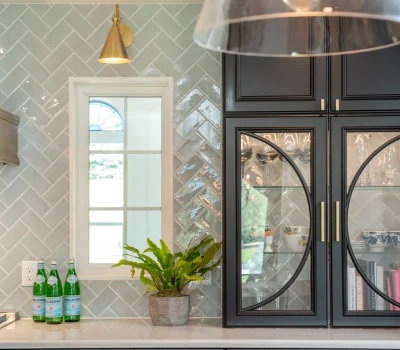 Modern kitchen with herringbone tile backsplash, black glass cabinets displaying glassware, potted plant, three bottles, and a gold wall lamp above.