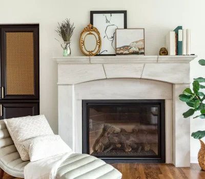 Stylish living room with fireplace, artwork, and books on mantel. Cozy chaise lounge, pillows, and leafy plant beside dark wooden cabinet.