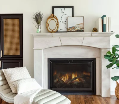 A cozy living room features a fireplace, decorative mantle items, lounge chair, large potted plant, and wooden cabinet with rattan details.