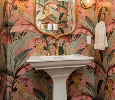 Decorative bathroom with vibrant tropical wallpaper, a pedestal sink, gold fixtures, and a rectangular mirror. Two lights illuminate the colorful space.