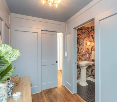 Bright hallway with light blue walls, wooden floor, decorative plant, and a vibrant, colorful bathroom visible through the open sliding door.
