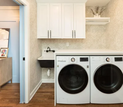 A laundry room with a washer, dryer, sink, and white cabinets. Adjacent room features a wooden dresser with framed artwork.