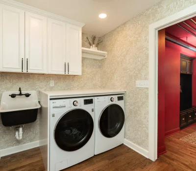 Laundry room with modern washer, dryer, black sink, white cabinets, and textured wallpaper. Adjacent red room with dark cabinetry visible.
