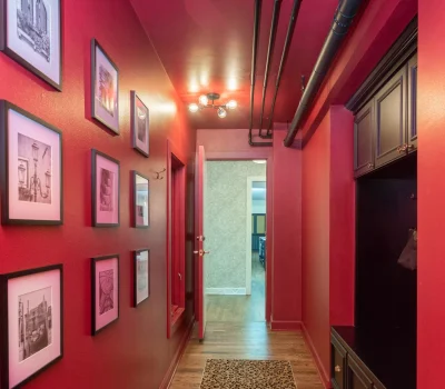 A vibrant red hallway with framed pictures, leopard print rug, and exposed ceiling pipes leading to an open door in a residential setting.