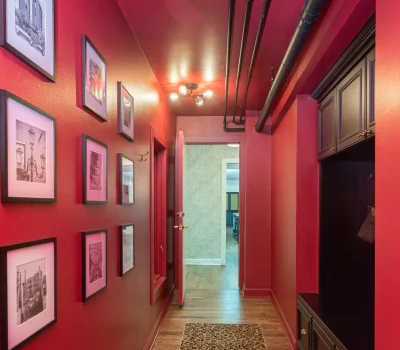 A narrow hallway with vibrant red walls features framed black-and-white art and a leopard-patterned rug, under exposed ceiling pipes.