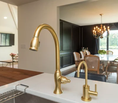 A modern kitchen with gold faucets overlooks a chic dining area featuring dark walls, elegant chandelier, and large windows with garden views.