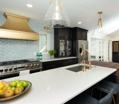Modern kitchen featuring a gold range hood, marble countertop, dark cabinets, herringbone backsplash, and elegant lighting fixtures, with fruit bowl centerpiece.