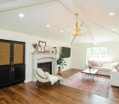Bright, spacious living room with white walls, wooden floor, elegant fireplace, chandelier, sectional sofa, and a large cabinet. Cozy, modern interior design.