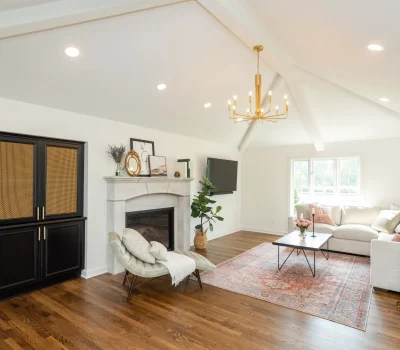 Bright living room with vaulted ceiling, elegant chandelier, cozy furniture, fireplace, and a decorative rug. A bar cart and indoor plants are present.