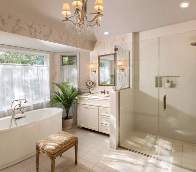 Elegant bathroom featuring a freestanding tub, glass shower, vanity with mirror, potted plants, and decorative chandelier under soft natural lighting.