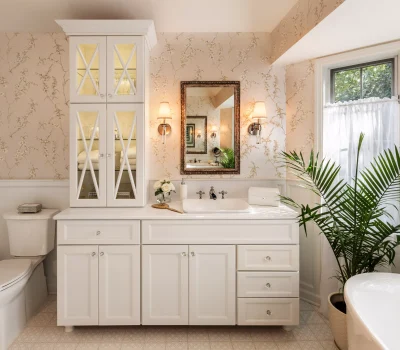 A bright bathroom features a vintage-style mirror, elegant wallpaper, white cabinetry, potted plant, and soft lighting, creating a cozy, classic atmosphere.