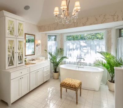 Bright bathroom with large bathtub, white cabinetry, patterned bench, chandelier, and potted plants. Light streams through sheer curtains, creating a tranquil atmosphere.