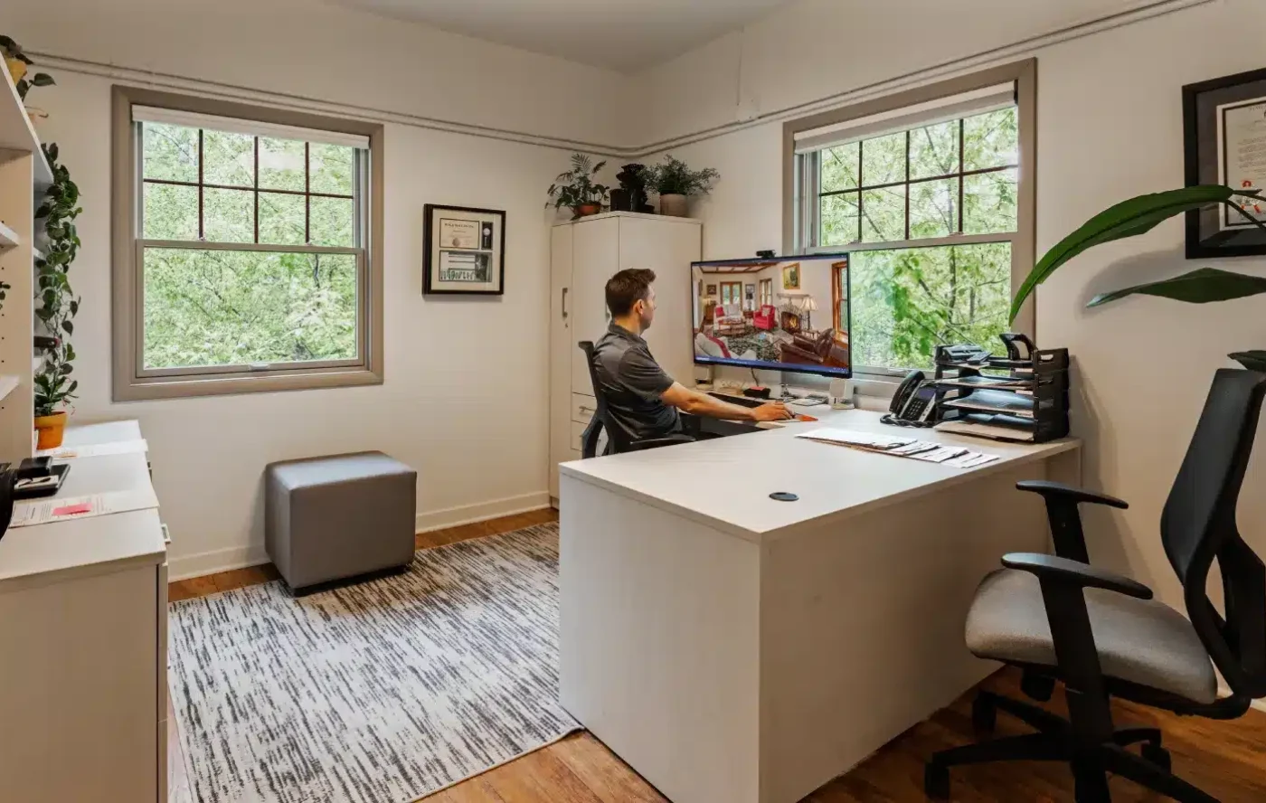 A person sits in a bright, modern home office with large windows, working at a computer. The room has plants and certificates.