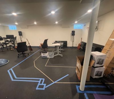 A spacious room with musical instruments, office chairs, and speakers. Boxes are stacked near a pillar; blue tape marks patterns on the floor.
