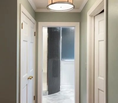 A hallway with light green walls, wooden floor, three doors, and a large round ceiling light fixture leading to a bathroom.