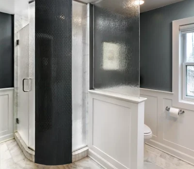 Modern bathroom with a curved glass shower, white paneled walls, and marble flooring. Natural light enters through a window, enhancing the sleek design.