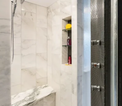 Luxurious white marble shower with built-in bench, wall niche storing toiletries, and modern stainless steel rain showerhead and controls. Bright lighting above.